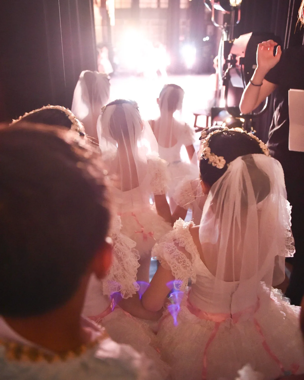 白いベールと花冠の衣裳の子どもたちが、舞台袖から光の差す舞台を見つめて出番を待つ後ろ姿